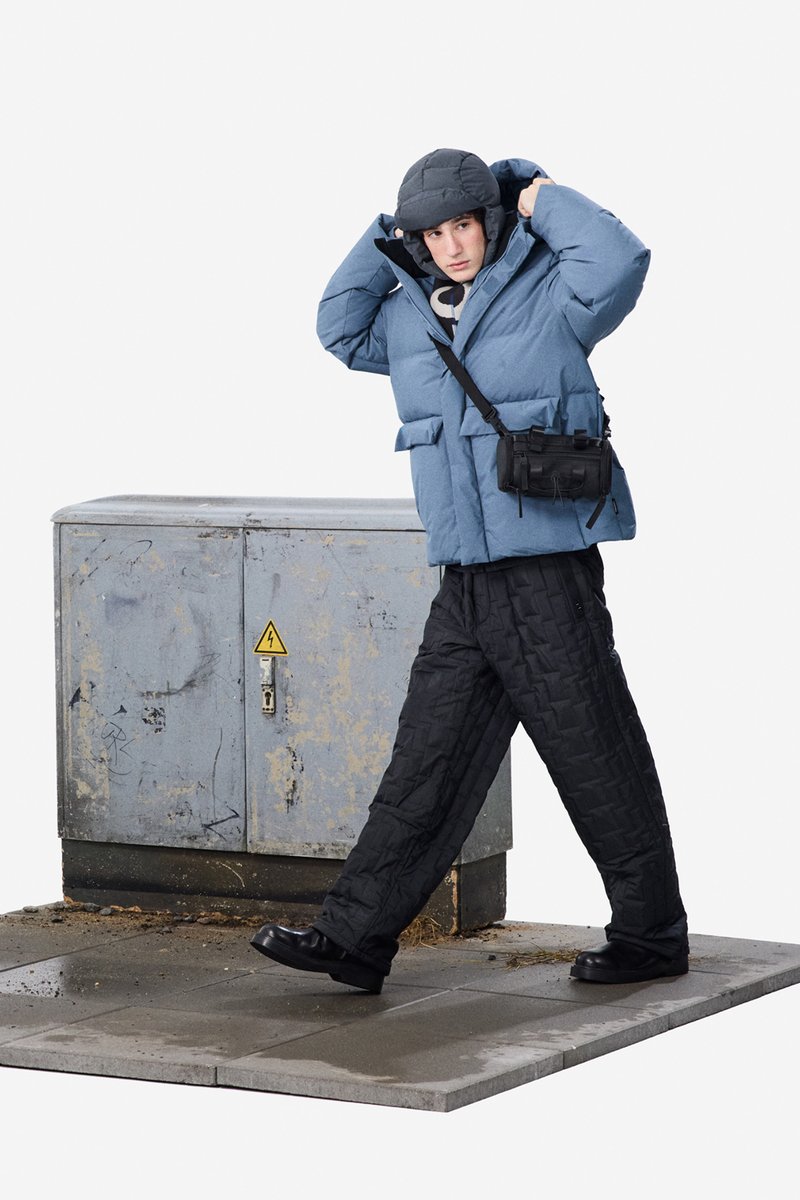 Gewatteerd blauwe jas, zwarte doorgestikte broek, platte zwarte schoenen en een grijze hoed. Zwarte crossbody tas. Staand op een grijs platform bij een nutsvoorzieningskast.