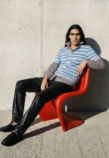 Man met lang donker haar die gelaagde gestreepte hemden en zwarte leren broeken draagt, leunt ontspannen in een rode moderne stoel tegen een betonnen muur.