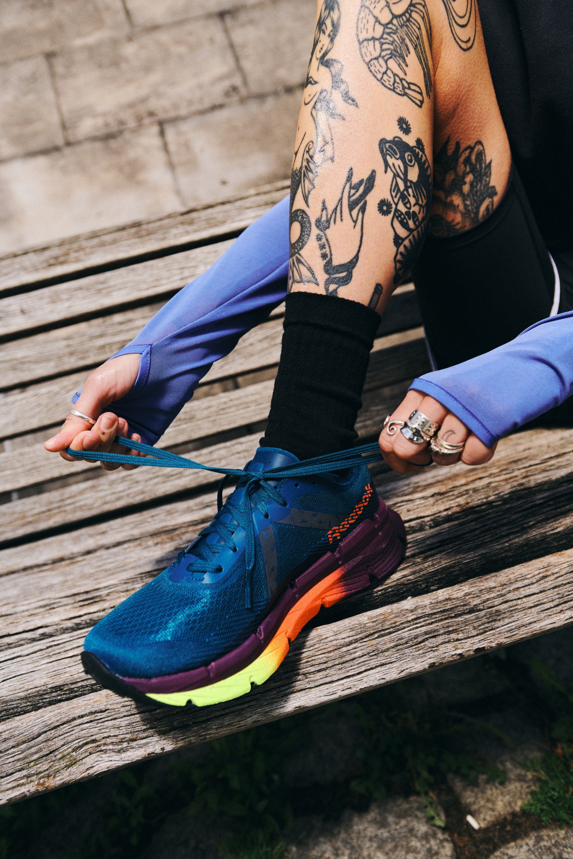 A tattooed arm holds bright blue shoelaces while tying a colorful running shoe, seated on a wooden bench in a sunny outdoor setting.