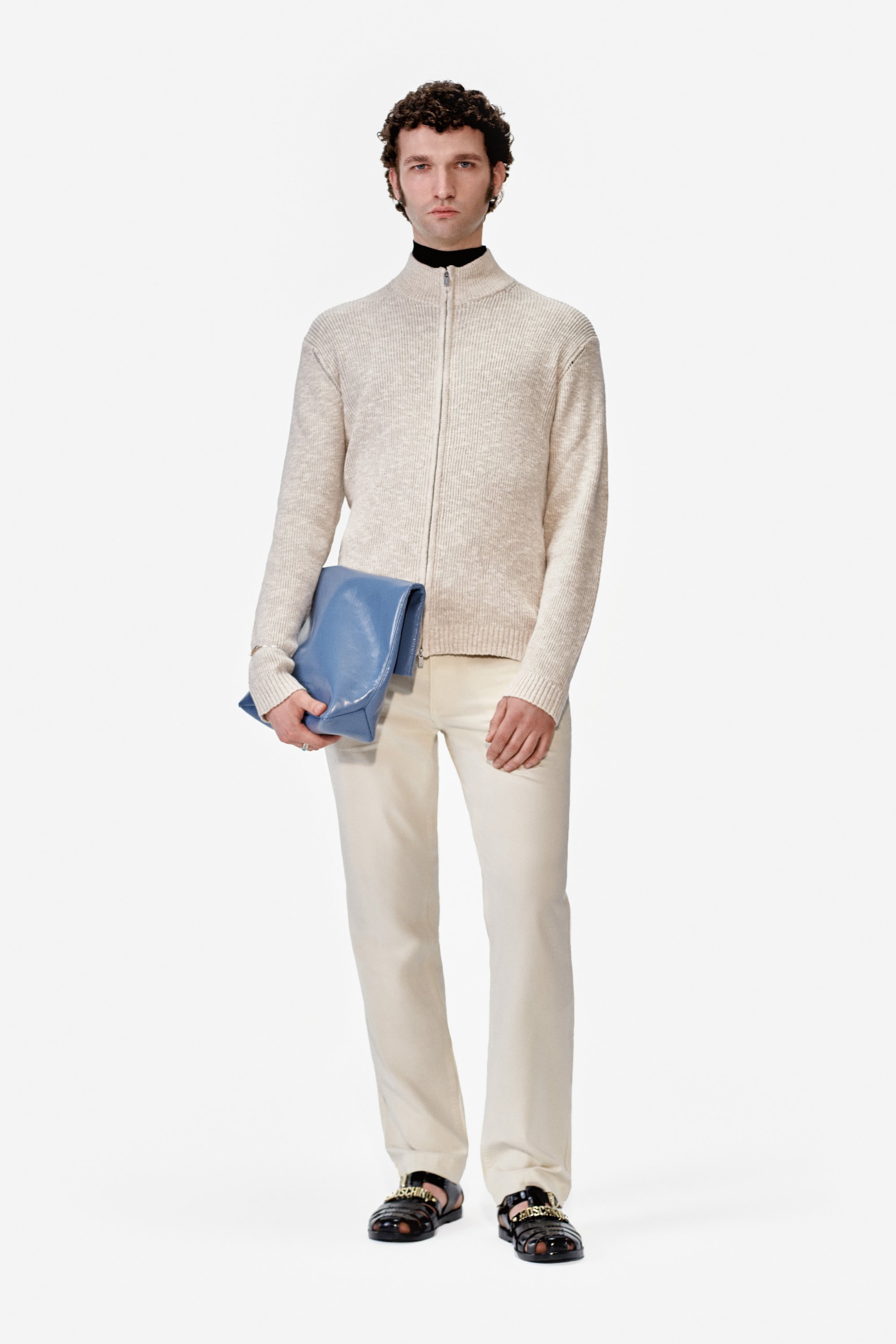 El modelo posa frente a un fondo blanco, vistiendo un suéter claro, pantalones color crema y sandalias negras, mientras sostiene un bolso de cuero azul.