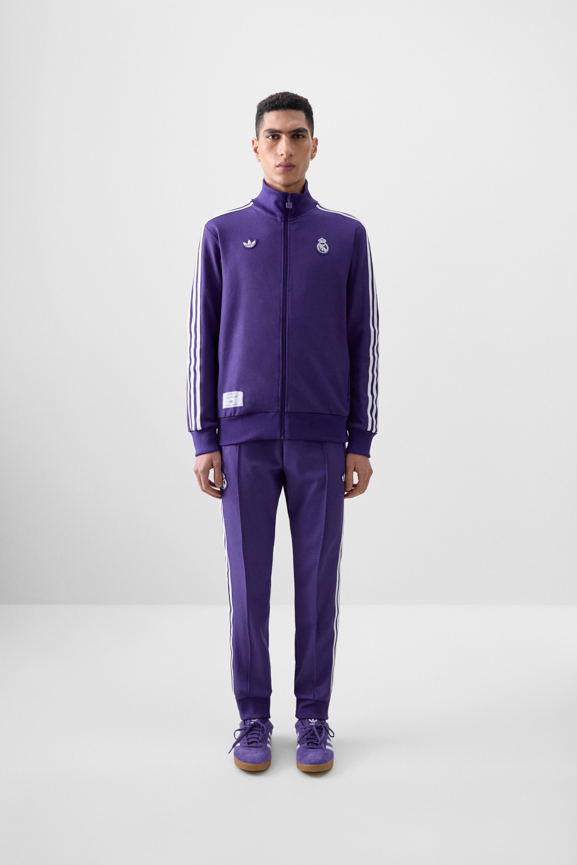 Een jonge man staat in een minimalistische omgeving, gekleed in een paarse trainingsoutfit met strepen en logo's, en maakt zijn look af met bijpassende schoenen.