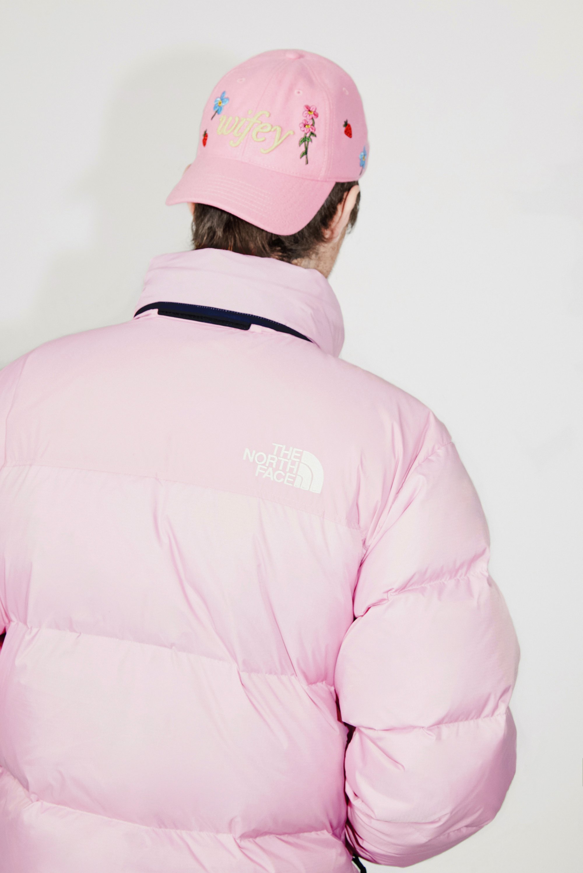 Una persona lleva puesta una chaqueta acolchada rosa y una gorra rosa con bordados florales y la palabra "wifey", de espaldas a la cámara.