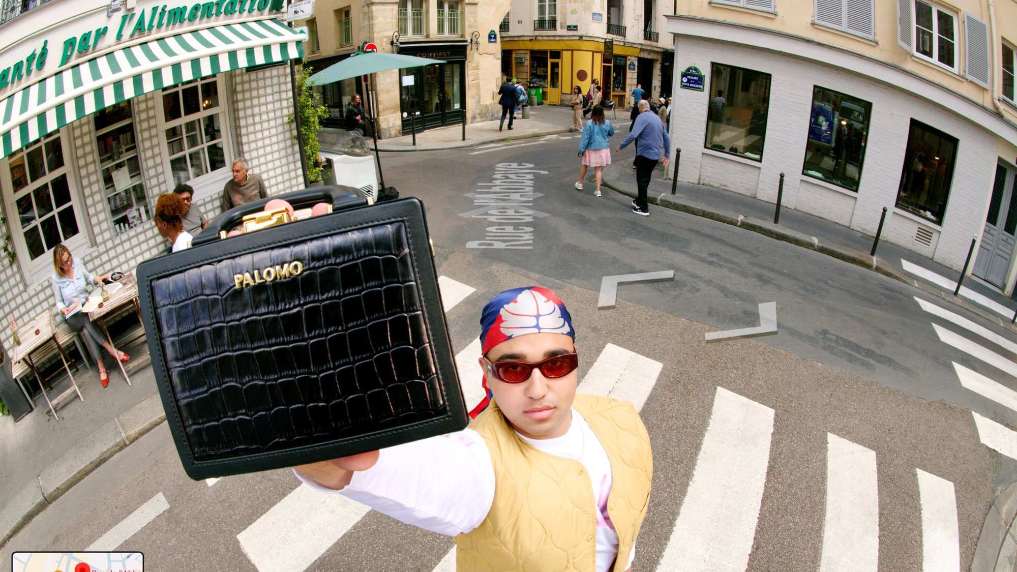 A person in sunglasses holds a black bag prominently while standing at a crosswalk near a café with patrons. Streets and buildings surround them.