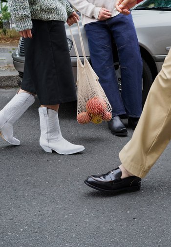 Four individuals stroll along a street, each wearing different types of footwear: white cowboy boots, black loafers, tan shoes, and blue wide-leg trousers. A net bag carries apples.