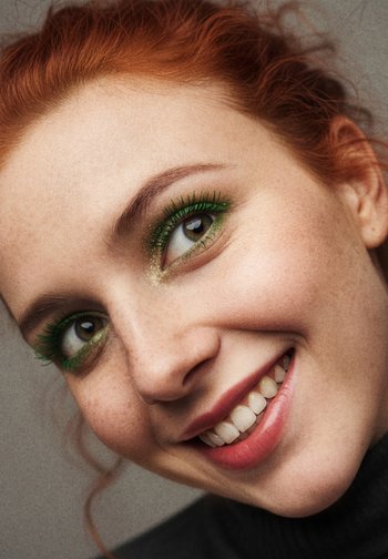 Close-up of a face with red hair. Features green eye makeup, prominent eyelashes, and a light pink lip color. Smooth skin with freckles.