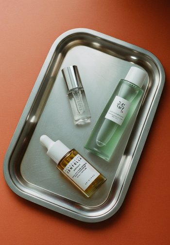 Three skincare bottles on a stainless steel tray: a clear glass bottle, a green liquid bottle, and an amber dropper bottle.