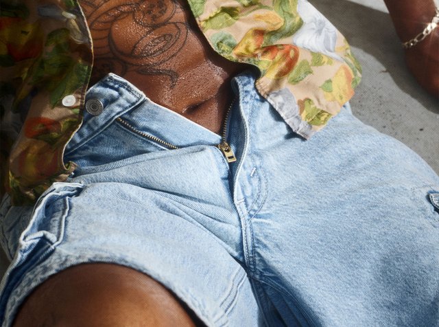 Personne musclée portant une chemise fleurie déboutonnée et un short en jean bleu clair avec la fermeture éclair partiellement ouverte, montrant une poitrine et un abdomen tatoués.