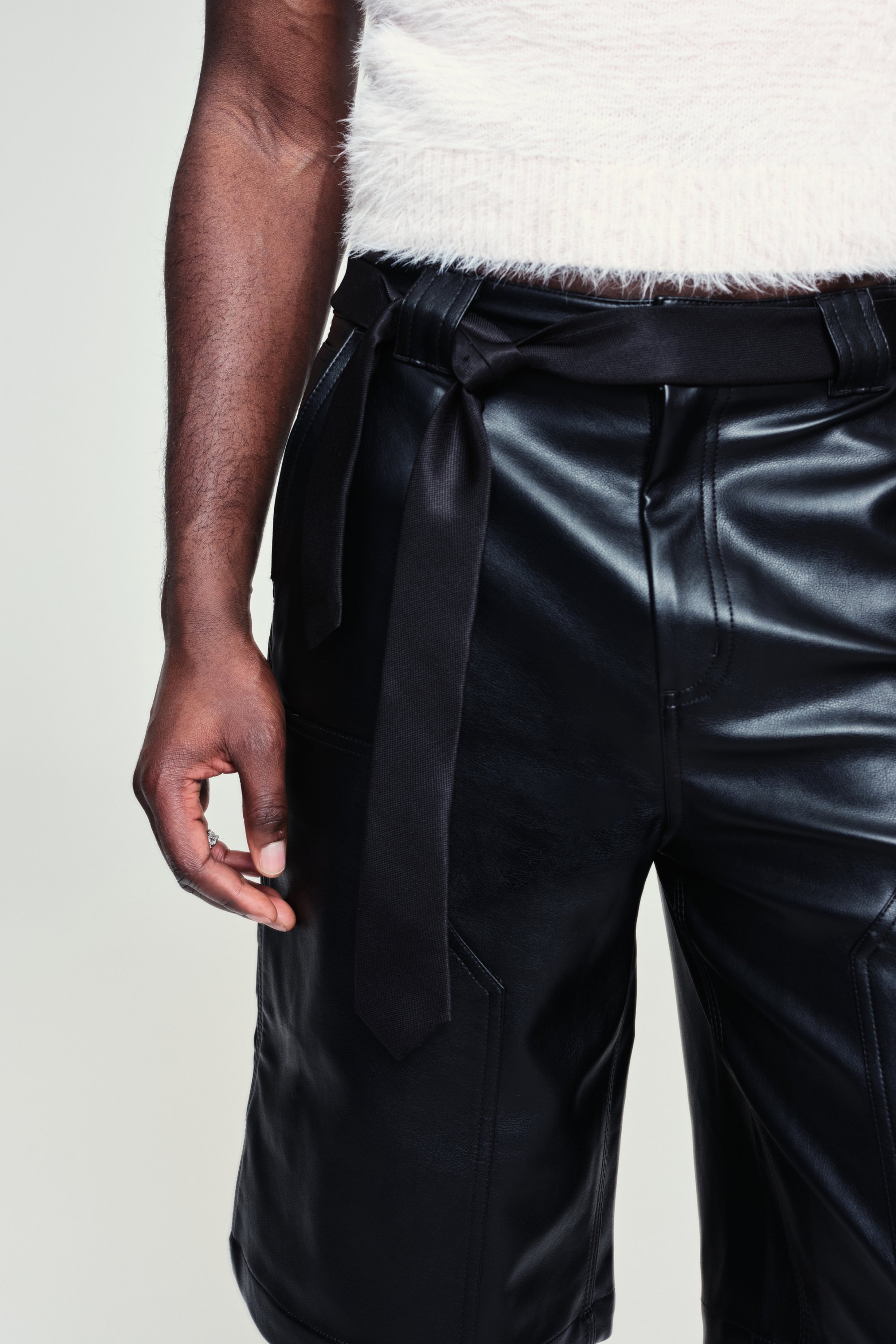 Primo piano di una mano che tiene dei pantaloncini neri in pelle con una vita legata, indossati con un top bianco morbido su uno sfondo neutro.