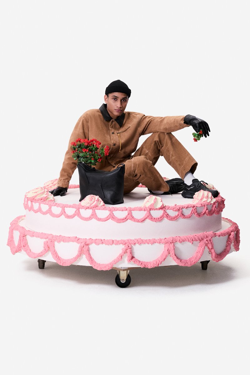 Persona con mono marrón y guantes negros sentada sobre un pastel de utilería de dos niveles rosa y blanco con flores rojas y un bolso negro encima.