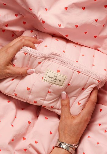 Ręce trzymają różową pikowaną saszetkę z czerwonymi sercami i etykietą z napisem The Flat Lay Co. London na pasującym, materiałowym tle w serduszka.