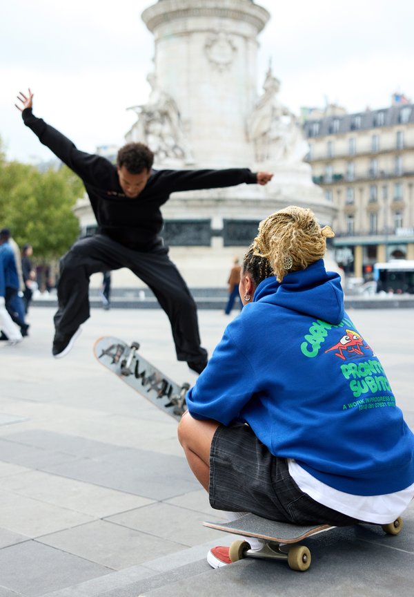 Skateboardere i en plass. En utfører et hopp i svarte bukser og en svart skjorte, mens en annen sitter i hettegenser i blått og shorts på en skateboard.