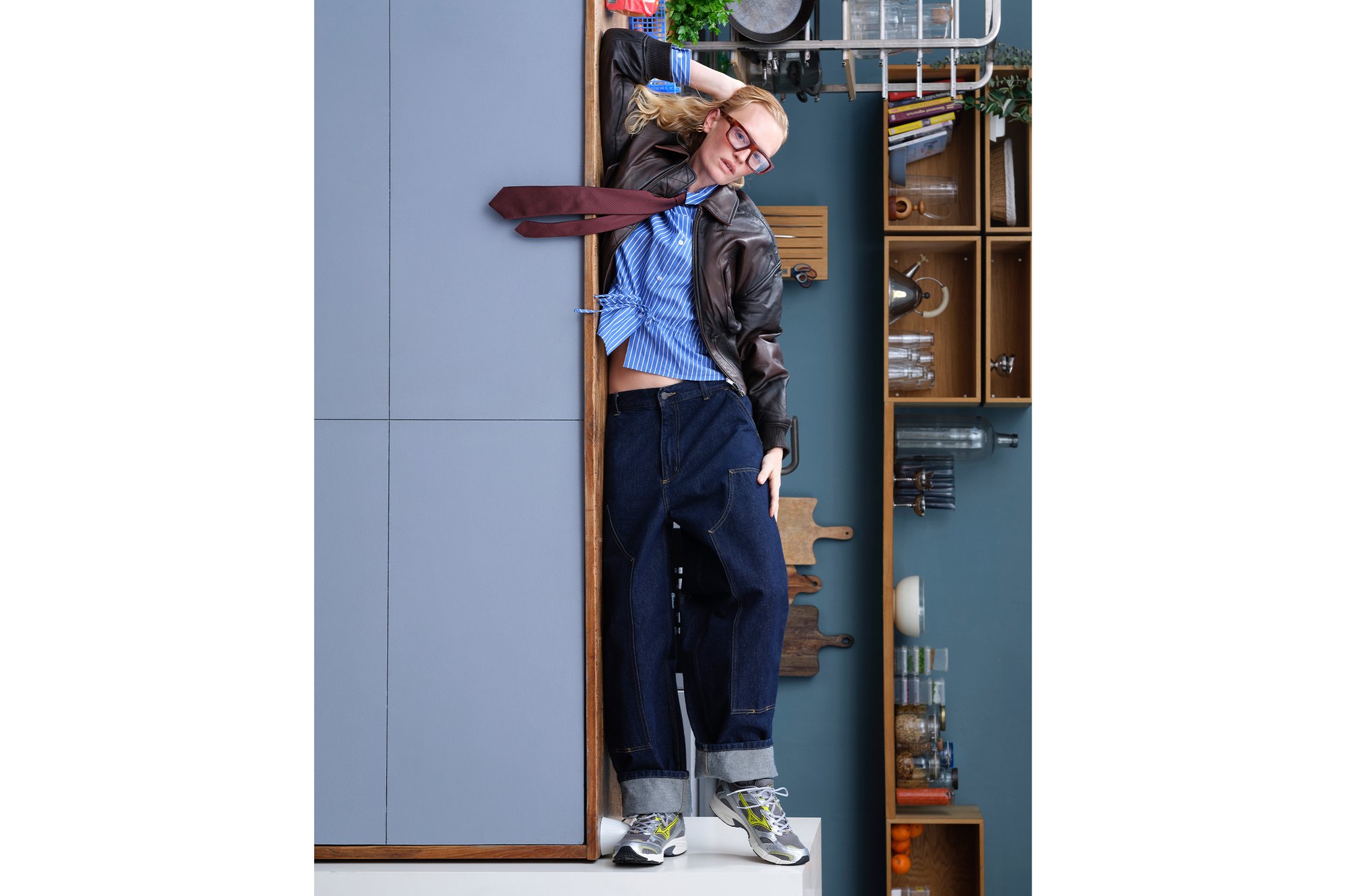 Una persona con pantalones de mezclilla oversized y una chaqueta de cuero se apoya de manera casual contra un armario de cocina, destacando una declaración de moda juguetona.