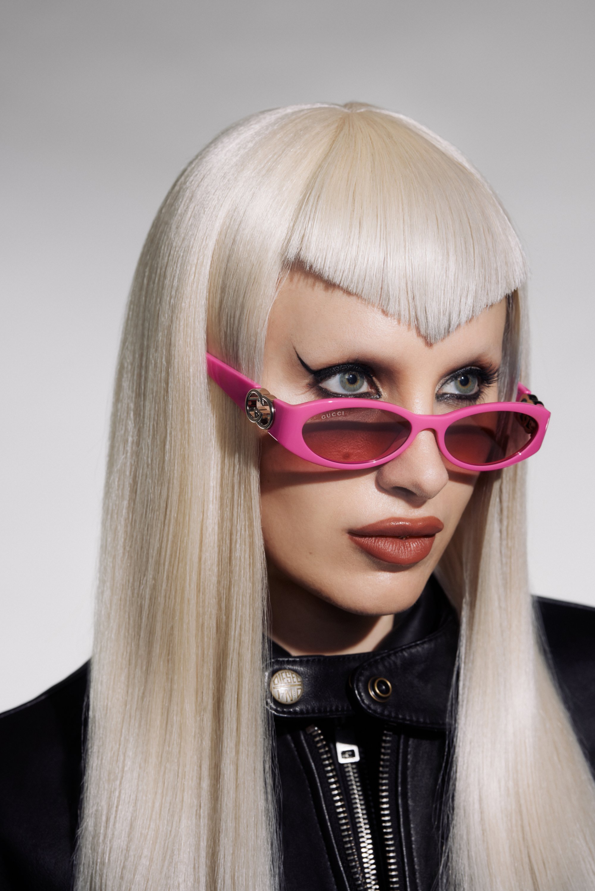 Modelo con cabello largo rubio y maquillaje audaz lleva gafas de sol rosas y una chaqueta de cuero negra, creando una impactante declaración de moda.
