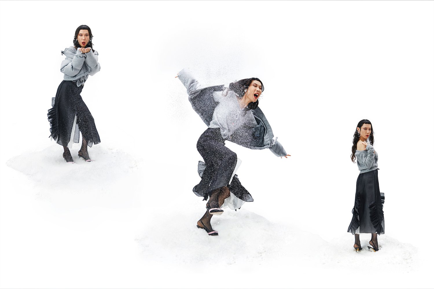 Jersey gris oversize, falda negra con volantes y tacones negros. La modelo posa sobre un fondo blanco nevado, creando un movimiento dinámico.
