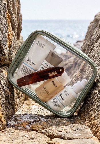 Durchsichtige Kosmetiktasche mit The Ordinary Hautpflege-Tuben und Spray, OUAI Shampoo und einem hölzernen Uppercut Deluxe Pflegekamm an felsiger Küste am Meer.