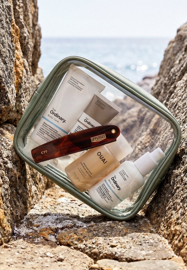 Durchsichtige Kosmetiktasche mit The Ordinary Hautpflege-Tuben und Spray, OUAI Shampoo und einem hölzernen Uppercut Deluxe Pflegekamm an felsiger Küste am Meer.