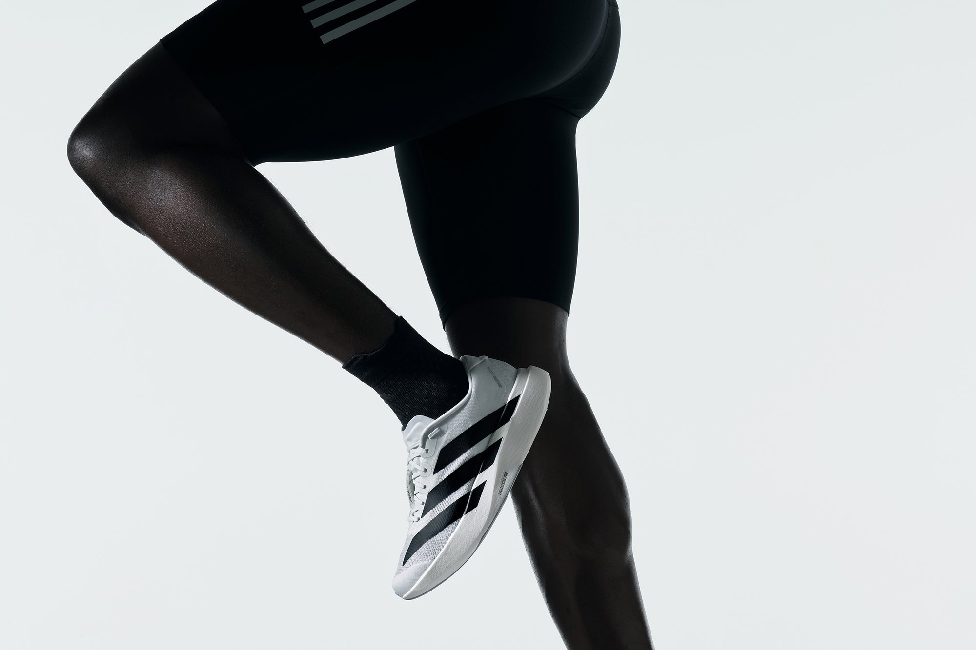 Un primer plano de una pierna musculosa en pantalones cortos deportivos negros y un calcetín negro, usando una zapatilla deportiva blanca con franjas negras, en medio de una zancada.