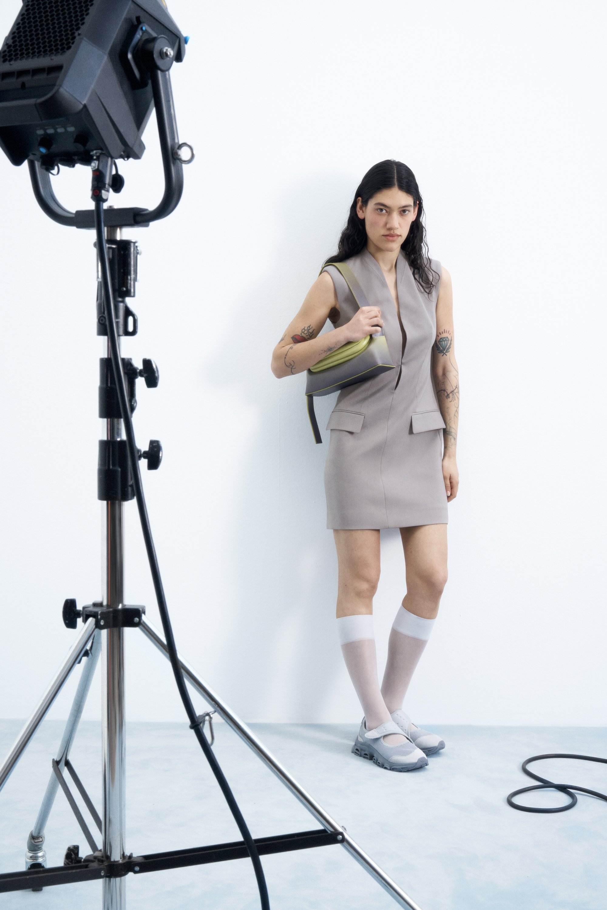 Modelo con un vestido gris ajustado sosteniendo un bolso verde, usando calcetas hasta la rodilla y zapatillas, se encuentra junto a un equipo de iluminación contra una pared blanca.