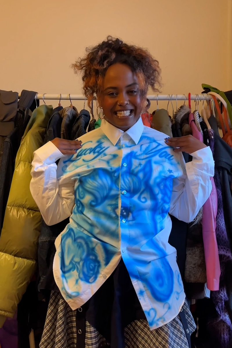 Een glimlachend persoon draagt een wit shirt met blauwe spray-patronen met de tekst "Lucky You", omringd door hangende kleding in een gezellige setting.