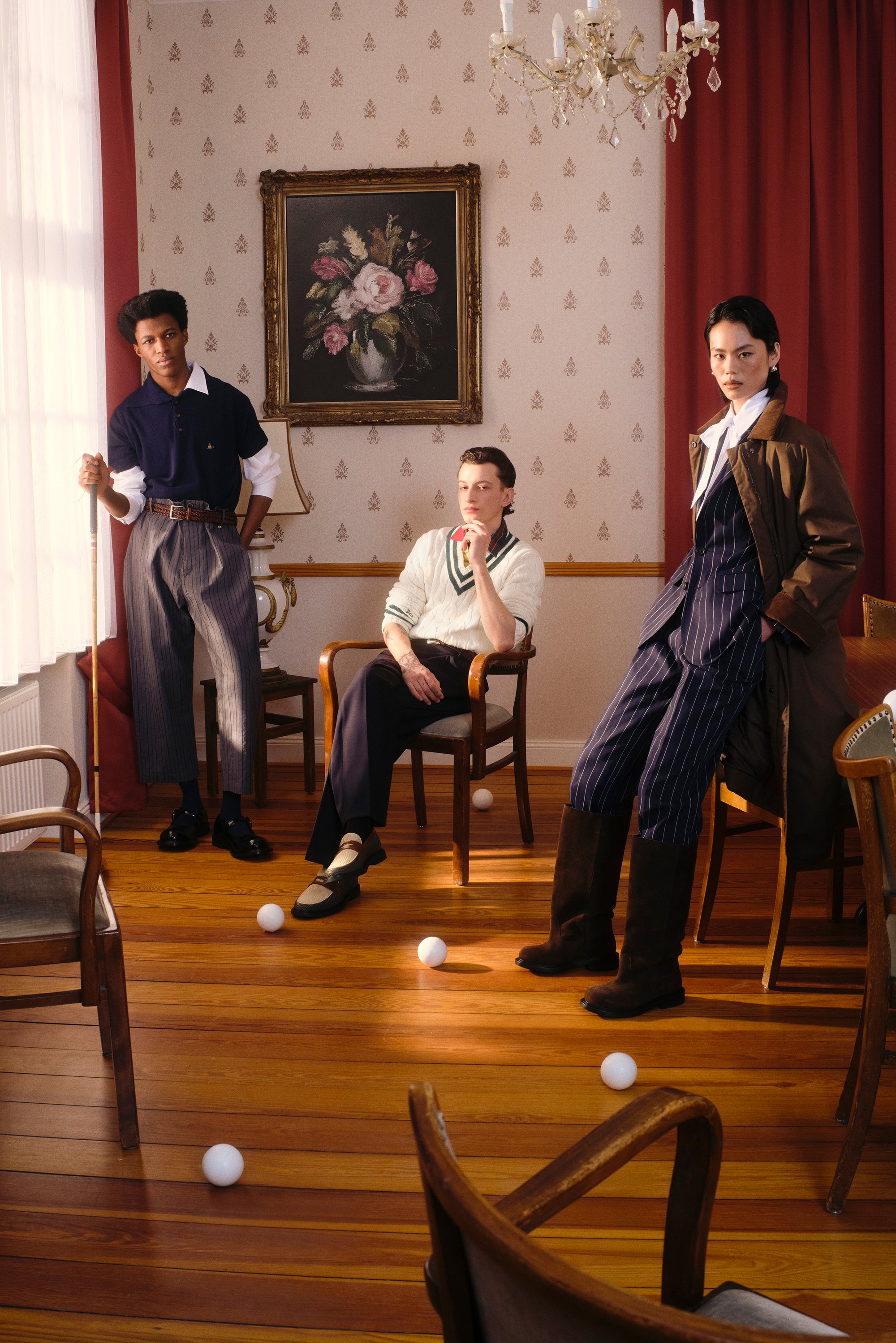 Tres personas elegantes posan en una habitación iluminada cálidamente con suelos de madera. Hay bolas blancas esparcidas por el suelo y una pintura floral adorna la pared.