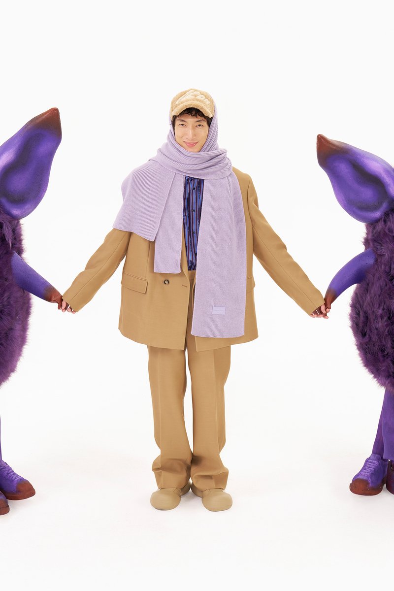 A person in a tan suit and purple scarf holds hands with two large, furry, purple characters with big eyes, set against a white background.