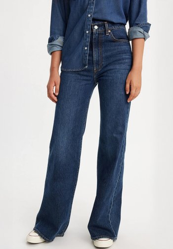 Falmede blå denim bukser med vide ben og høy midje, frontlommer og lett slitasje. Kombinert med en denim skjorte med knapper. Hvite sneakers.