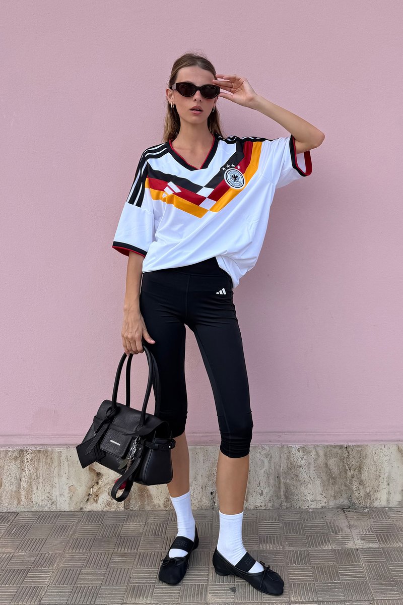 Frau trägt ein Deutschland Fußballtrikot, schwarze Leggings, weiße Socken, schwarze Schuhe, Sonnenbrille, hält eine schwarze Handtasche, steht vor einer pinken Wand.