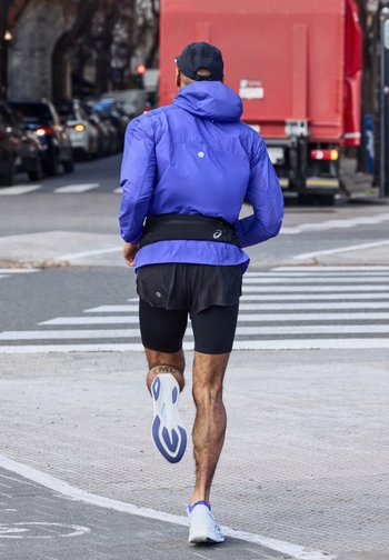 Δρομέας με μπλε μπουφάν και μαύρο σορτς τρέχει σε δρόμο της πόλης κοντά σε διάβαση πεζών, με αυτοκίνητα και ένα κόκκινο φορτηγό στο φόντο.