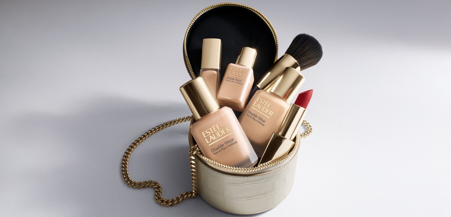 Beigefarbene runde Lederhandtasche mit goldener Kettenriemen, die Estée Lauder Foundation-Flaschen, einen Make-up-Pinsel und roten Lippenstift auf einer weißen Oberfläche hält.