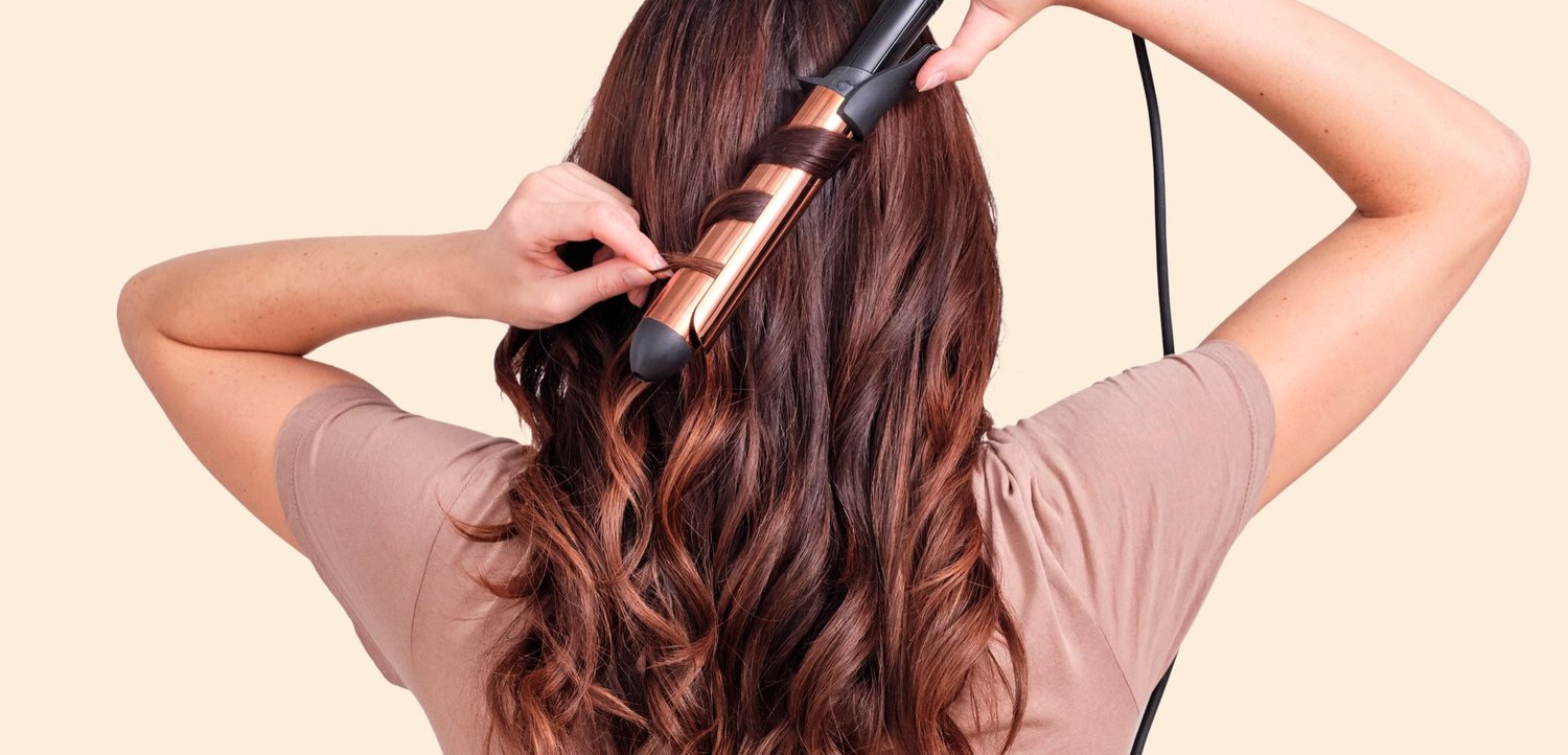 Person lockt lange braune Haare mit einem kupferfarbenen Lockenstab, von hinten gegen einen hellen Hintergrund gesehen.