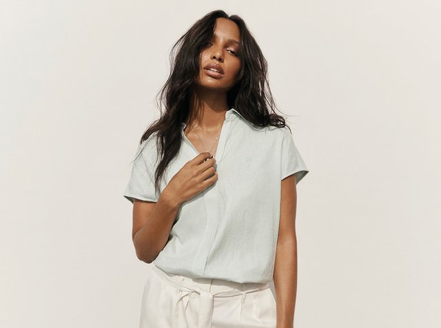 Femme aux longs cheveux foncés portant un chemisier léger à manches courtes et un pantalon blanc, tenant une main près du col sur un fond uni.