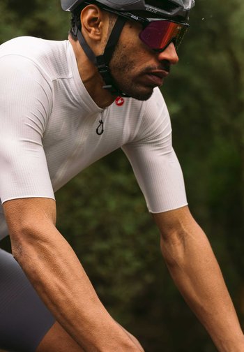 A focused cyclist in a fitted white jersey and helmet, showcasing muscular arms, rides against a blurred green background.