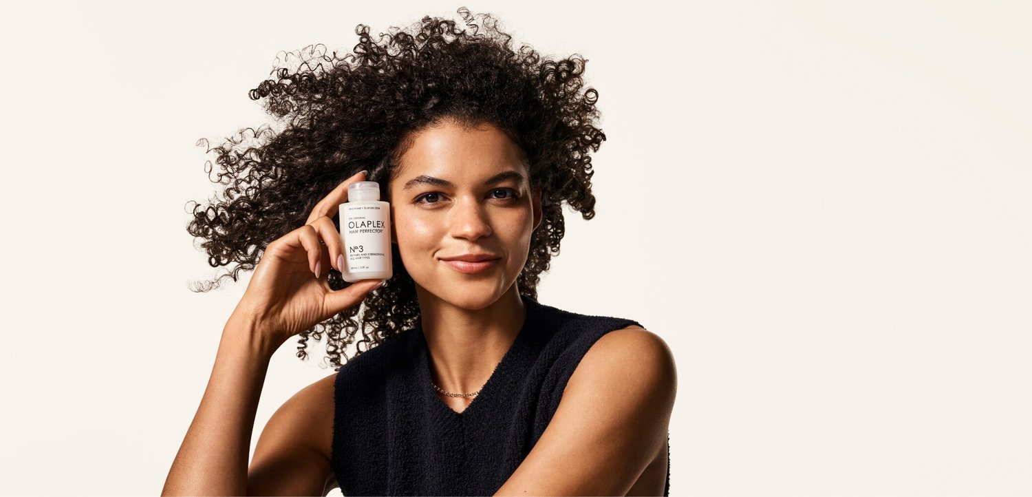 Donna con capelli ricci che tiene vicino al viso una piccola bottiglia bianca di Olaplex No. 3 hair perfector, sorridendo, su uno sfondo chiaro e uniforme.