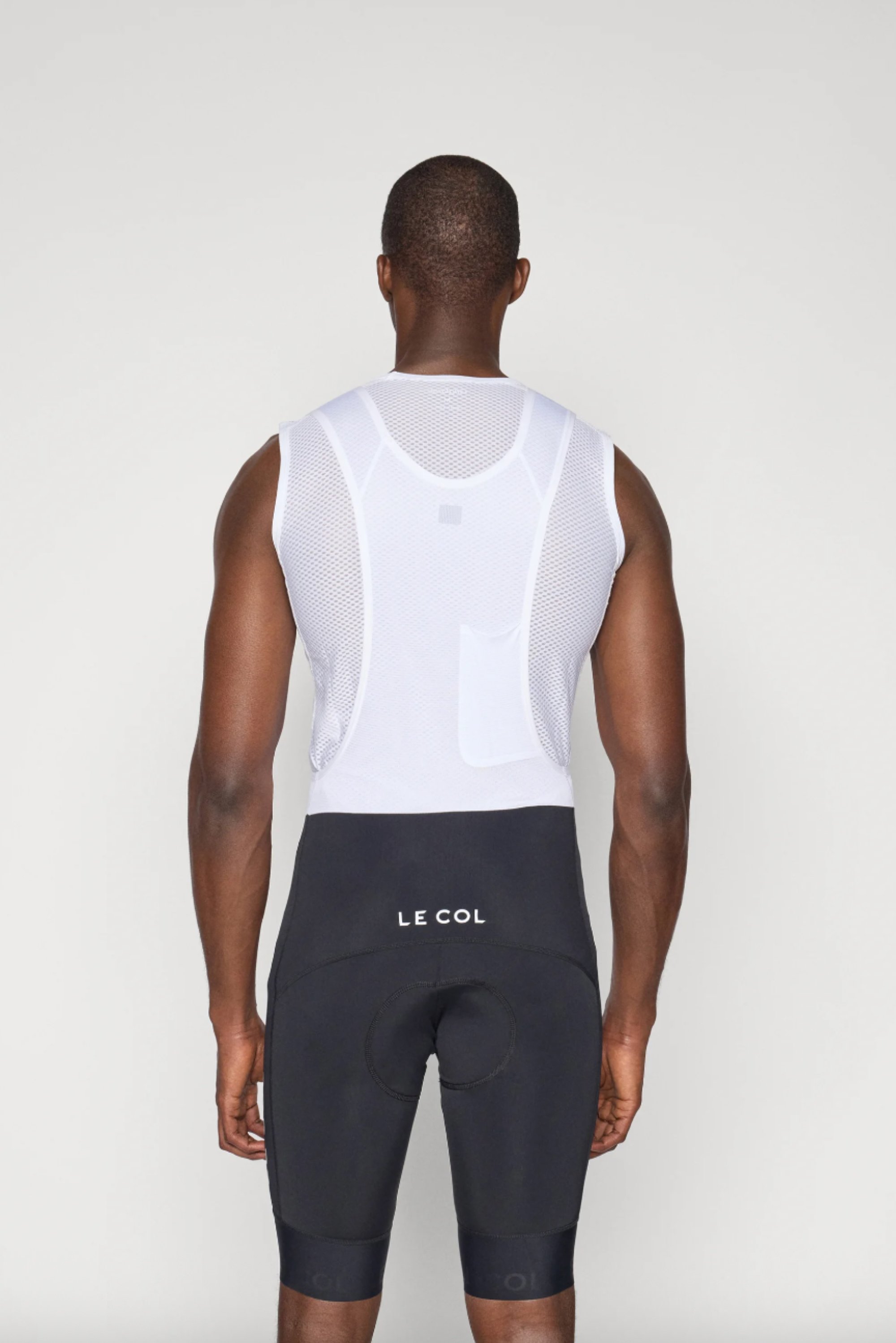 Eine Person steht mit dem Rücken zur Kamera, trägt ein weißes ärmelloses Trikot und schwarze Radshorts mit "LE COL"-Logo.