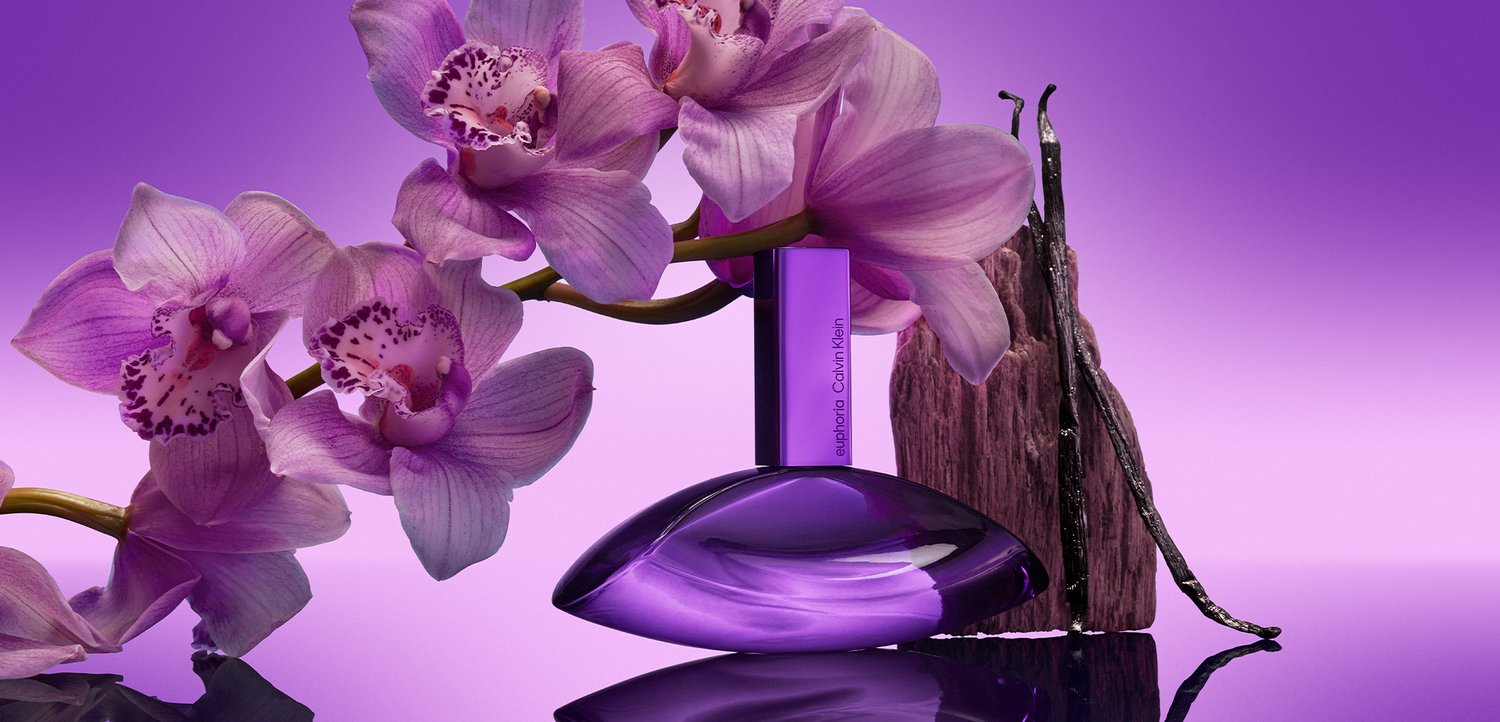 Lilla orkidéblomster ved siden af en lilla Calvin Klein-parfume flaske og vaniljebælge på en reflekterende overflade med lilla baggrund.