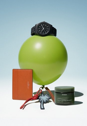 Schwarze Armbanduhr auf grünem Ballon mit Lederkartenetui, Palmen-Schlüsselanhänger und Glas mit Aveda Feuchtigkeitscreme, arrangiert auf weißer Oberfläche.