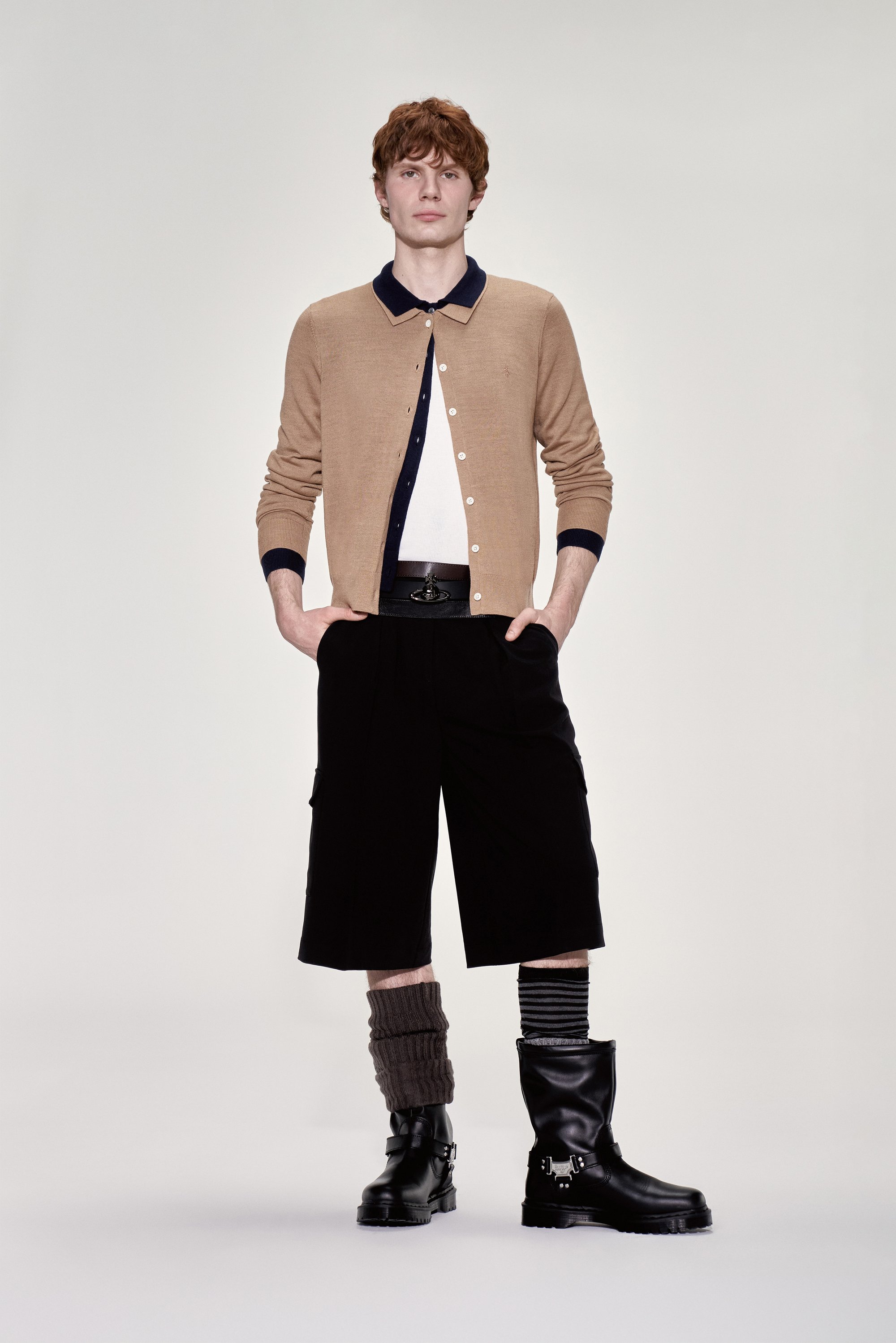 Un joven se encuentra de pie con confianza, llevando un cárdigan color beige sobre una camisa blanca, combinado con pantalones cortos negros y botas negras gruesas.