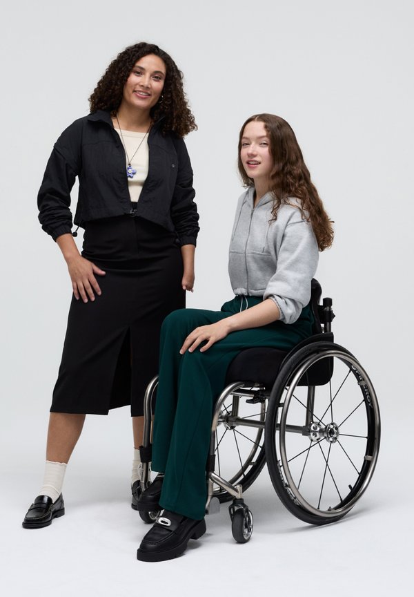 Due donne, una in giacca nera corta e gonna, l'altra in felpa grigia e pantaloni verdi su una sedia a rotelle, entrambe con mocassini.