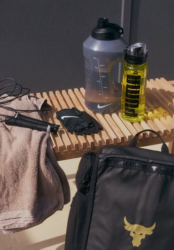 Un sac à dos noir avec un logo doré, une bouteille d'eau transparente, une serviette de sport grise, des gants d'entraînement noirs et une corde à sauter sur une surface en bois.