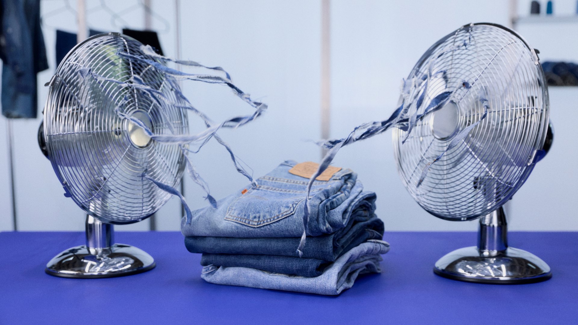Twee draaiende ventilatoren blazen strengen rafelige denim naar een stapel opgevouwen jeans op een blauwe tafel, met kleerhangers waarop meer denim wordt getoond op de achtergrond.