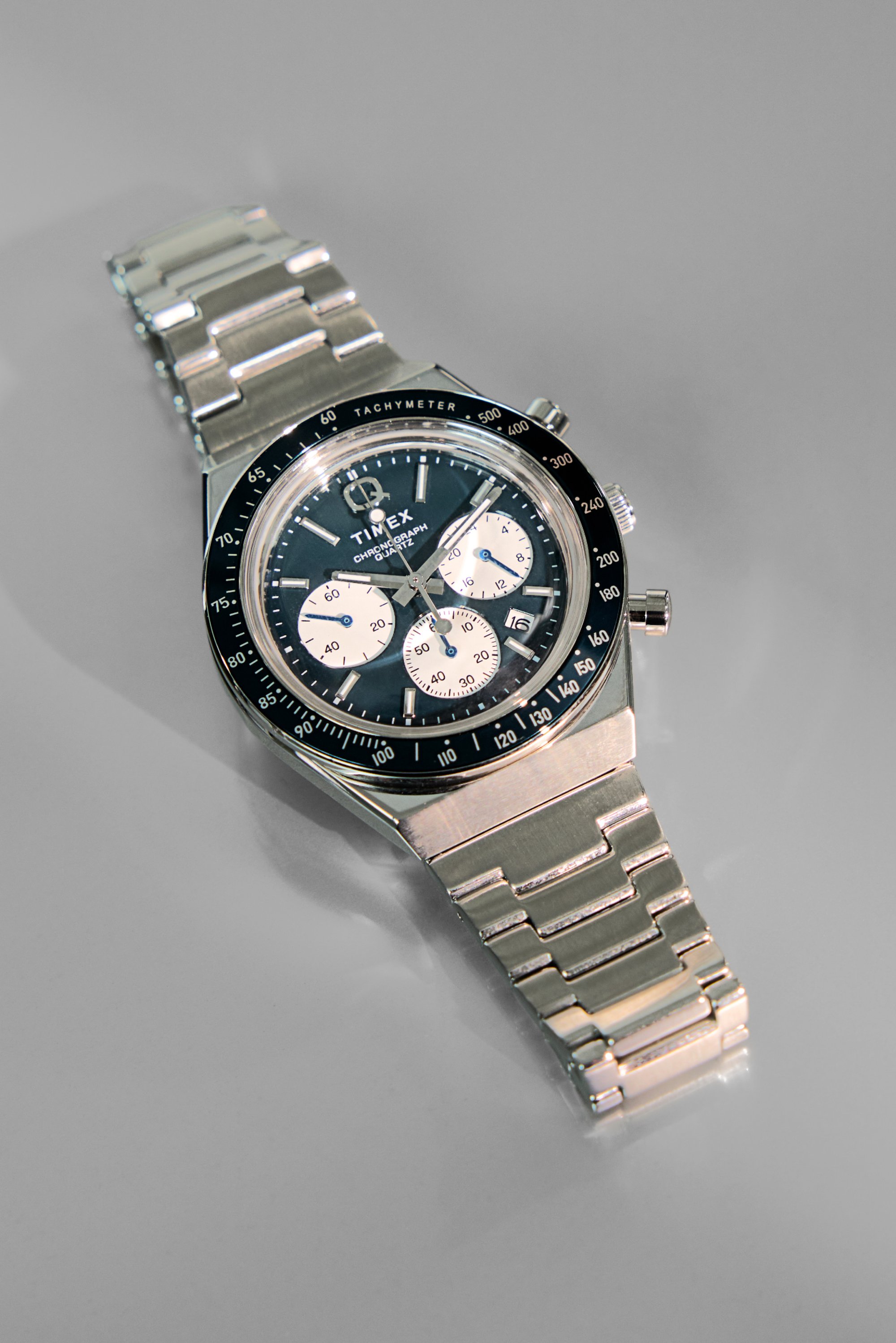 Reloj cronógrafo de acero inoxidable plateado con esfera negra, tres subesferas blancas, bisel taquímetro y pulsera de eslabones. Ventana de fecha mostrada.