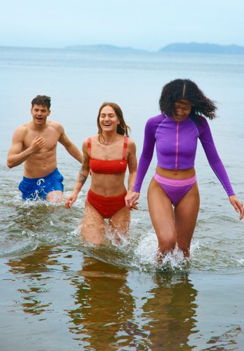 Tre unge voksne smiler og løper ut av grunt vann på en strand, to kvinner i badedrakter og en mann i badebukser.