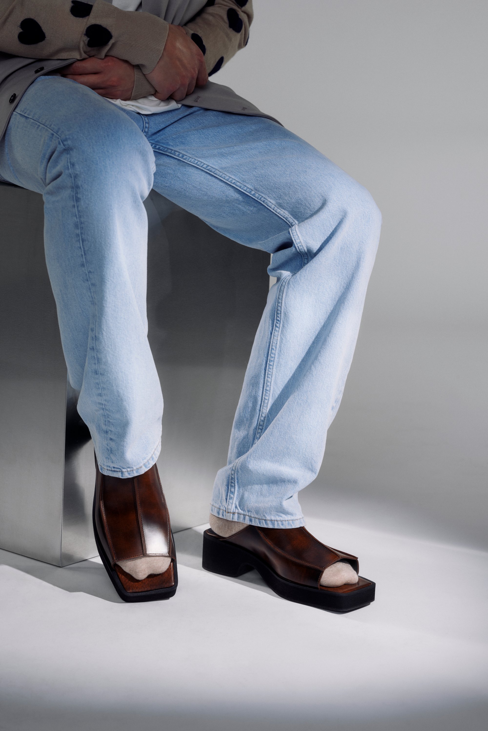 Persoon zittend op een metalen oppervlak, gekleed in blauwe jeans en een beige trui, met stijlvolle bruine open-tipped schoenen met een stevige hak.