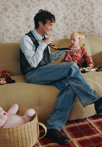 Barn som leker med leker på en teksturert gul sofa. En voksen i svart vest og blå skjorte holder en leke. Brunt tregulv med et mønstret teppe i nærheten.
