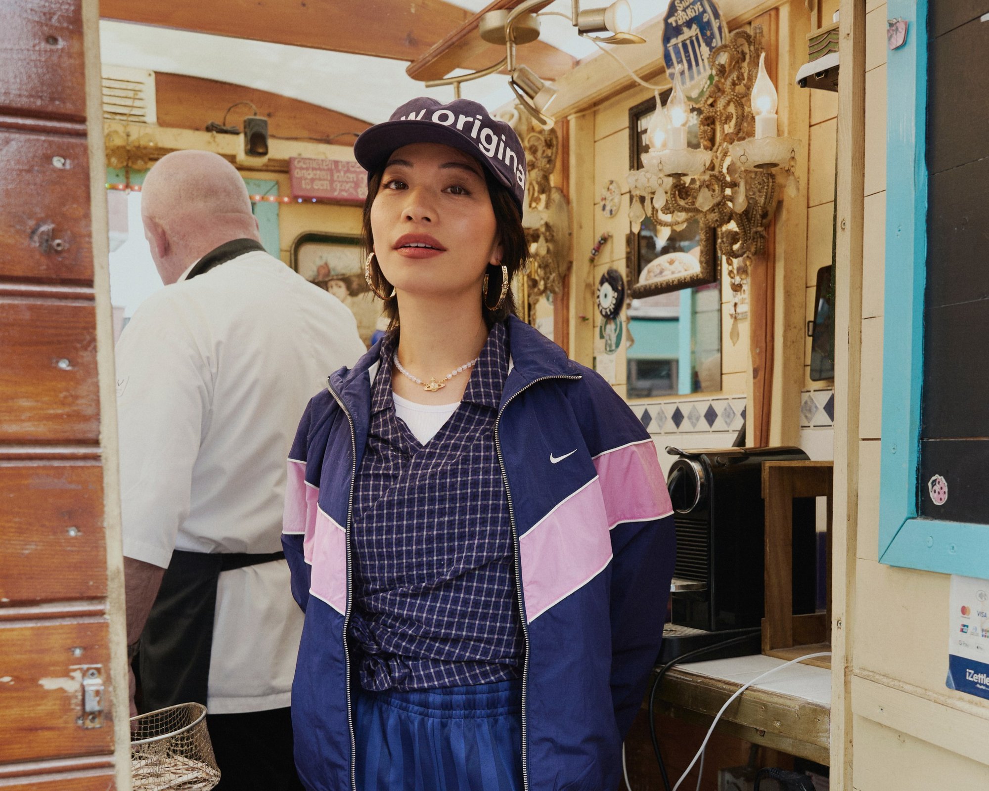 Veste Nike marine et rose sur un haut à carreaux, associée à un pantalon bleu. Les accessoires comprennent une casquette de baseball et des boucles d'oreilles créoles. Fond en bois.