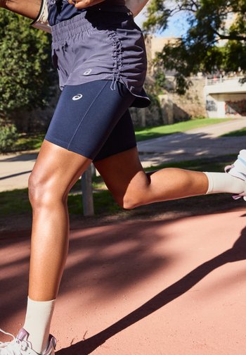 Άτομο τρέχει σε εξωτερικό χώρο φορώντας σκούρο μπλε σορτς και μωβ φούστα, με λευκές κάλτσες και ροζ παπούτσια τρεξίματος με λεπτομέρειες, σε μια ηλιόλουστη μέρα.