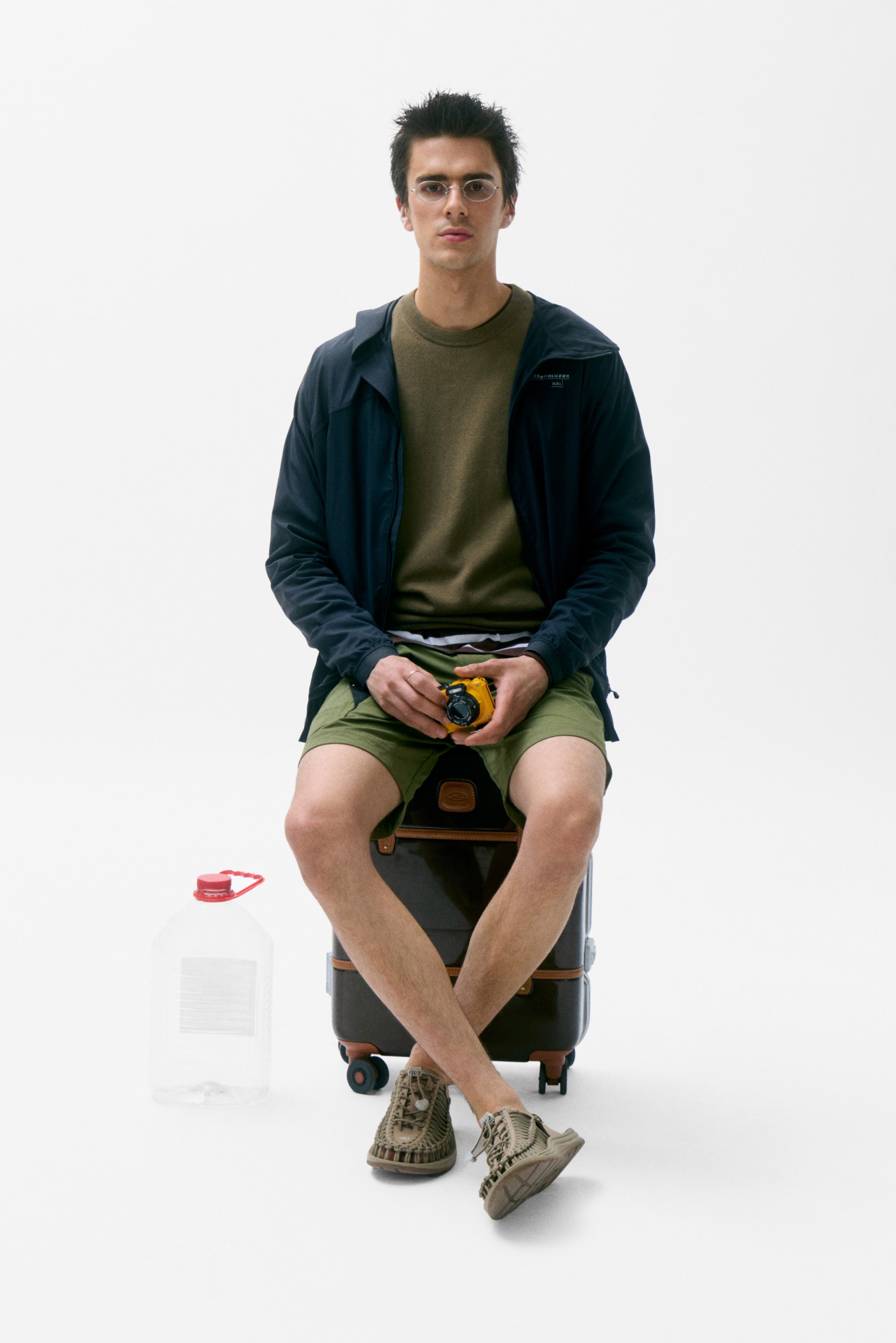 Un joven con gafas está sentado sobre una maleta, sosteniendo una cámara amarilla. Lleva una chaqueta oscura, una camiseta verde, pantalones cortos y zapatos informales. Hay una garrafa de agua cerca.
