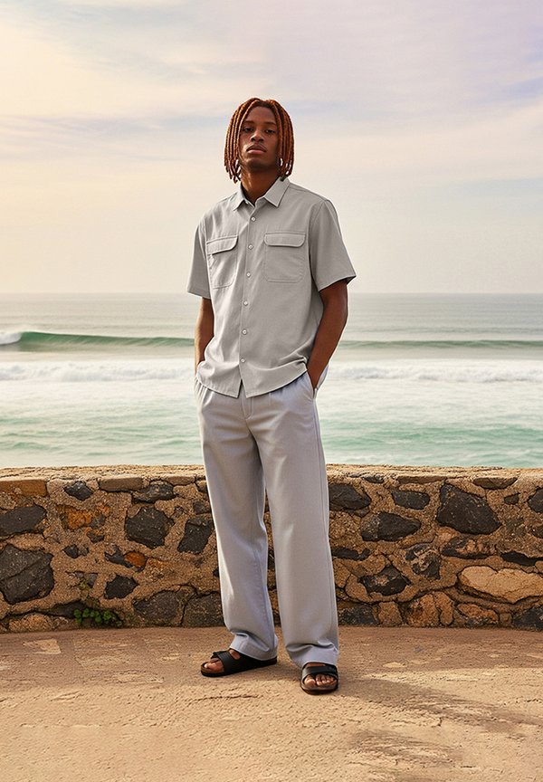 Light grey short-sleeve shirt with two chest pockets, matched with light grey wide-legged trousers and black sandals, set against a coastal backdrop.