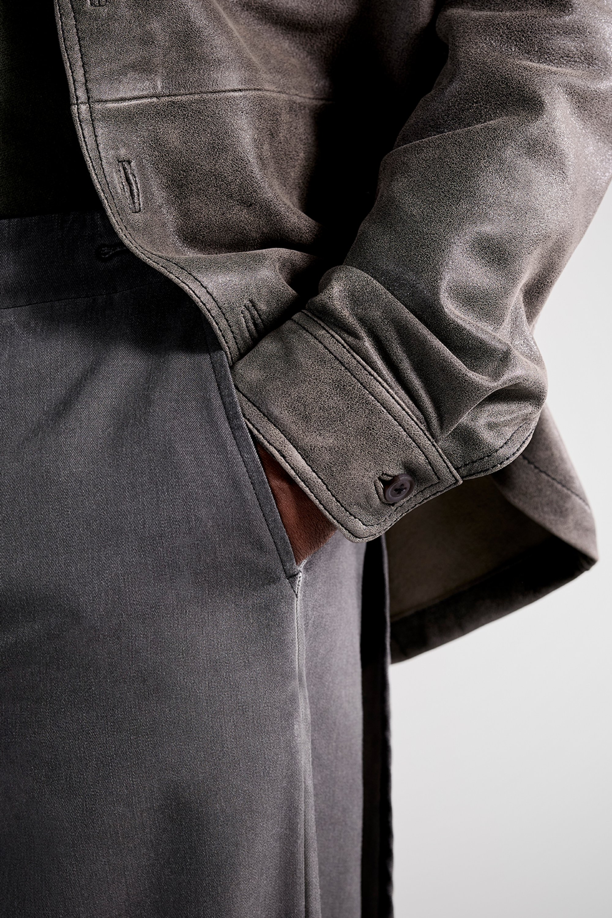 A close-up of a hand resting in the pocket of dark trousers, partially covered by a slate-colored leather jacket. The fabric texture is visible.
