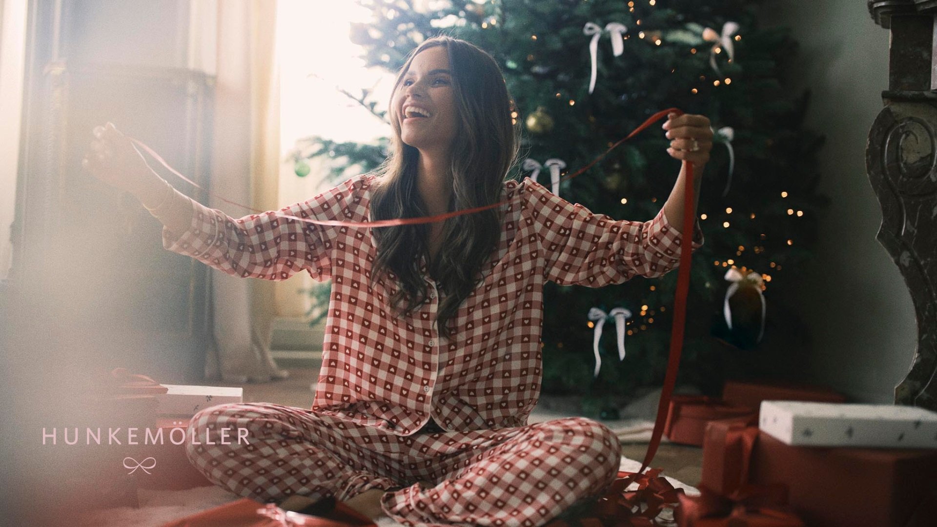 Vrouw in rood geruite pyjama die naast een versierde kerstboom zit, glimlacht en houdt een rood lint vast met ingepakte cadeaus in de buurt.